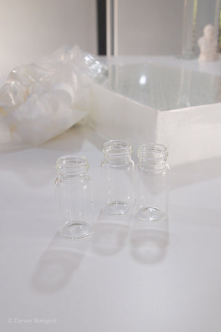 Three small clear glass vials with screw-top necks arranged on a white surface, with a white box and a crumpled translucent plastic bag in the softly blurred background.