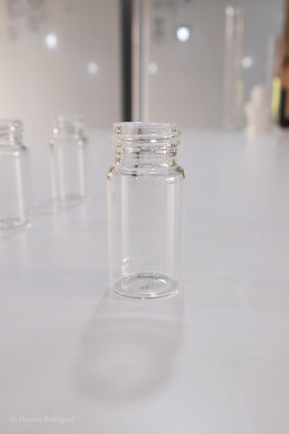 A small clear glass vial with a threaded neck, standing on a white surface with other similar vials blurred in the background.