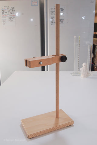 A wooden scientific single retort stand with a rectangular base, vertical support rod, and horizontal arm holding a black clamp, shown on a white tabletop with a blurred laboratory background.