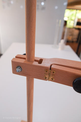 Close-up of a wooden adjustable single retort stand or frame component with a light natural wood finish, brass hinge, and black knob, set against a softly blurred indoor background.